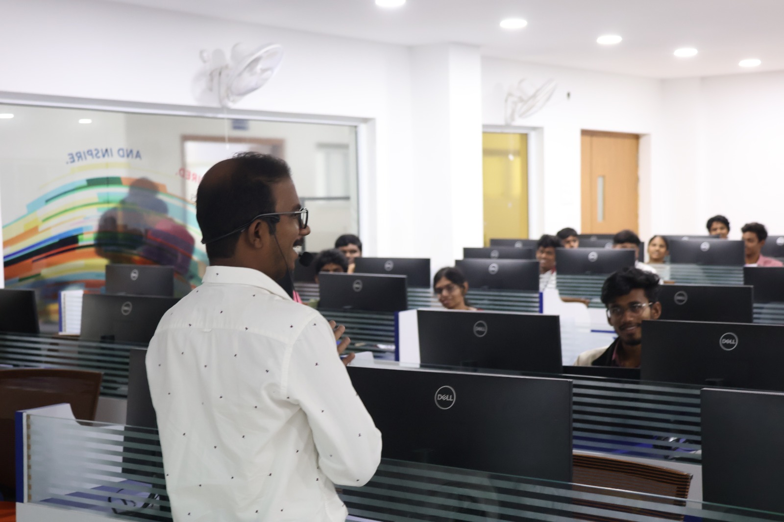 A TechM4India mentor addressing smiling students seated behind rows of monitors in a computer lab